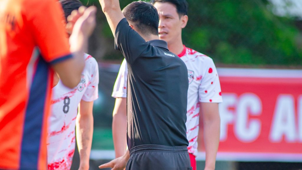 ผู้ตัดสินสวมชุดดำยกใบเหลืองขึ้นเหนือศีรษะระหว่างการแข่งขันฟุตบอล มีผู้เล่นสวมเสื้อส้มและเสื้อลายจุดแดงยืนล้อมอยู่