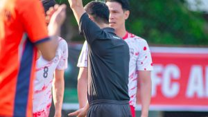 ผู้ตัดสินสวมชุดดำยกใบเหลืองขึ้นเหนือศีรษะระหว่างการแข่งขันฟุตบอล มีผู้เล่นสวมเสื้อส้มและเสื้อลายจุดแดงยืนล้อมอยู่