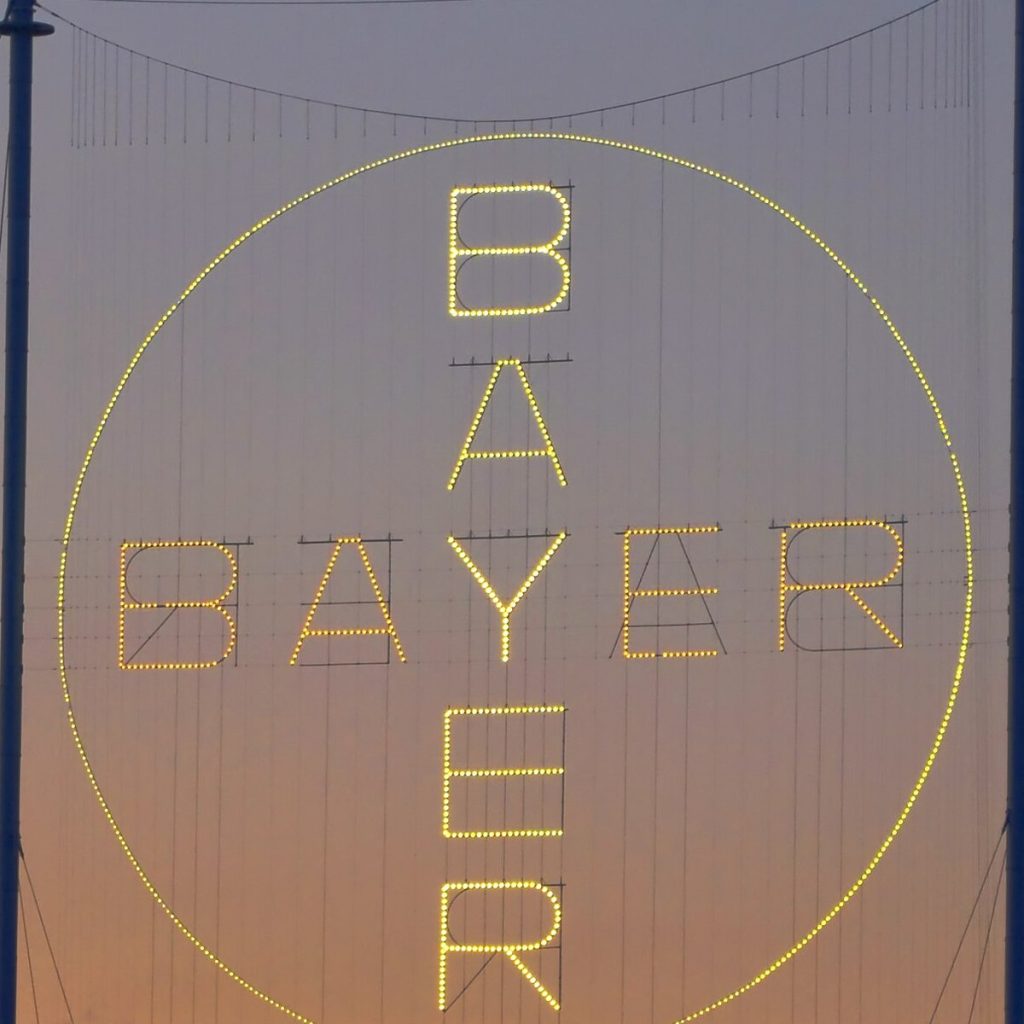 ภาพระยะใกล้ของป้ายไฟวงกลมสะกดคำว่า BAYER เห็นดวงไฟทุกดวงชัดเจนบนพื้นหลังท้องฟ้ายามเย็น