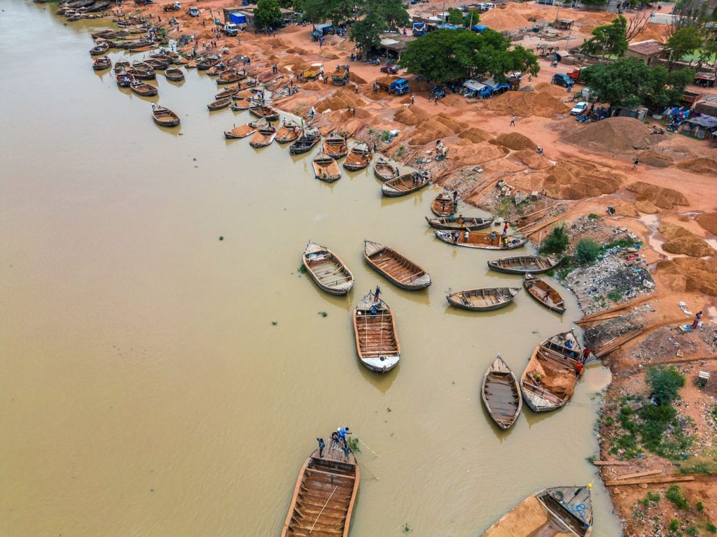 ภาพมุมสูงของเรือไม้ลำเล็กจำนวนมากจอดและแล่นอยู่บนแม่น้ำสีน้ำตาล ริมฝั่งเป็นกองทรายและผู้คนทำงานอยู่ตามแนวตลิ่ง
