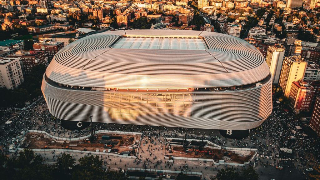 ภาพมุมสูงของสนามซานติอาโก เบร์นาเบวยามพลบค่ำ เห็นหลังคาโค้งสีขาวรอบสนามและตึกอาคารที่พักอาศัยโดยรอบในกรุงมาดริด