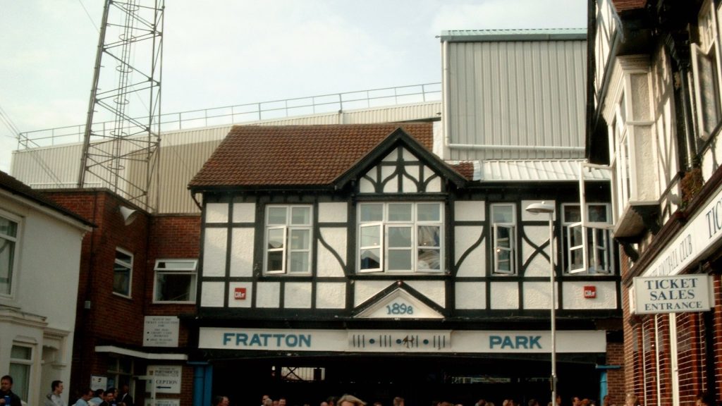 อาคารทางเข้าแบบทิวดอร์สีขาวดำของสนามฟรัตตัน พาร์ค มีป้ายชื่อ FRATTON PARK และปีก่อตั้ง 1898 อยู่เหนือทางเข้า