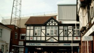 อาคารทางเข้าแบบทิวดอร์สีขาวดำของสนามฟรัตตัน พาร์ค มีป้ายชื่อ FRATTON PARK และปีก่อตั้ง 1898 อยู่เหนือทางเข้า