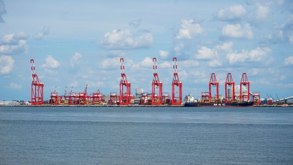 เครนขนถ่ายตู้คอนเทนเนอร์สีแดงขาวเรียงรายที่ท่าเรือลิเวอร์พูล มีเรือสินค้าจอดเทียบท่า