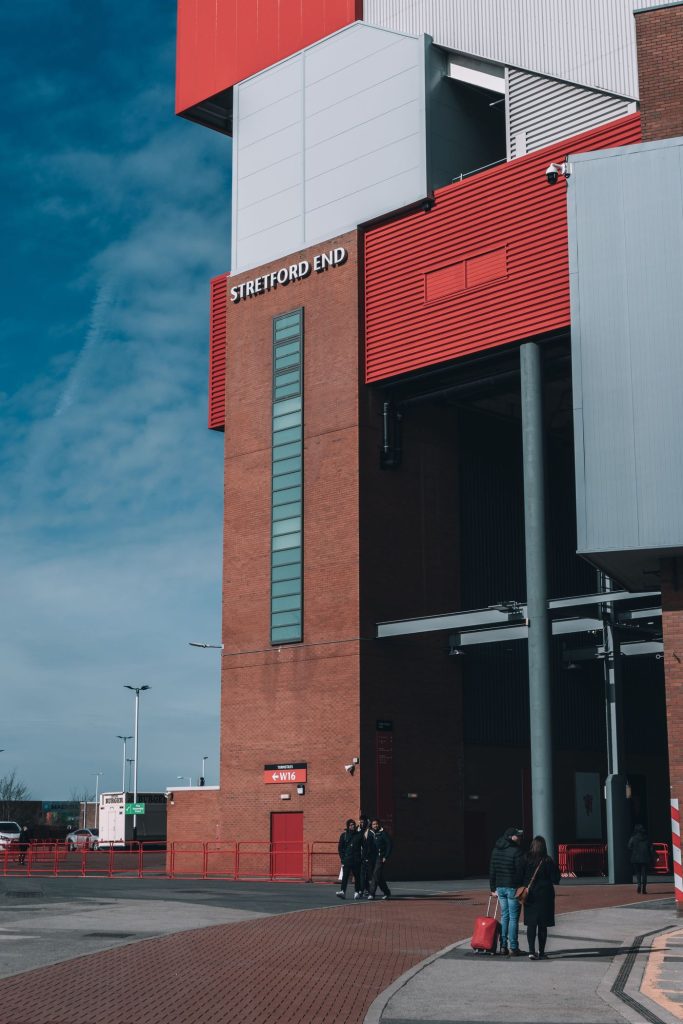 ด้านนอกอาคารสเตรตฟอร์ด เอนด์ ของสนามโอลด์ แทรฟฟอร์ด มีป้ายชื่อ STRETFORD END ติดอยู่บนผนังอิฐสีแดง และมีผู้คนเดินอยู่ด้านหน้าทางเข้า